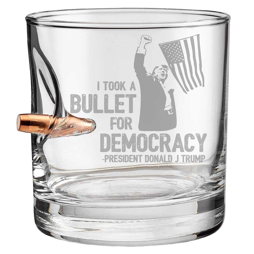 Trump whiskey glass featuring a bullet design and the phrase "I took a bullet for democracy" with a flag graphic.